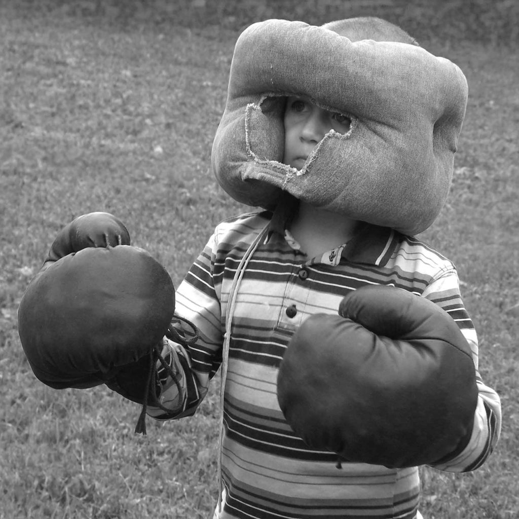 Black and white photo of small boy wearing striped shirt with oversized boxing gloves and headgear