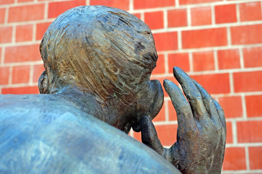 "Bronze statue of a person leaning forward with a hand to their ear, appearing to listen intently, set against a red brick wall background."